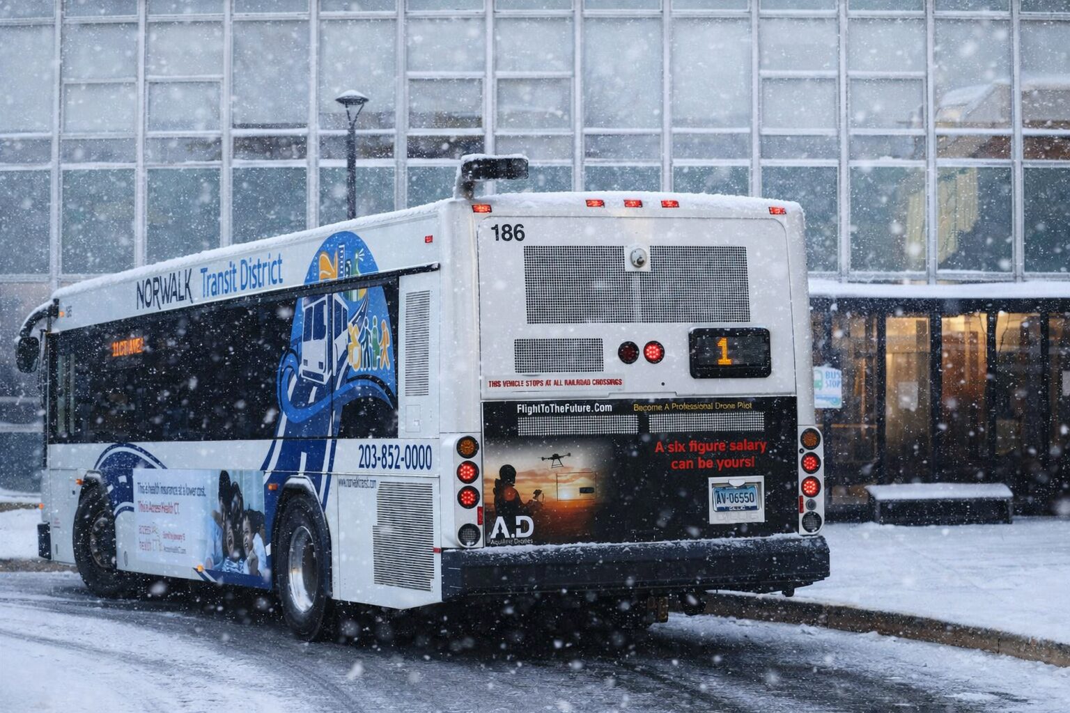 A Norwalk Transit District bus parked at a curb in winter conditions, light snow falling, with a glass building in the background and route number 1 displayed on the back of the bus.